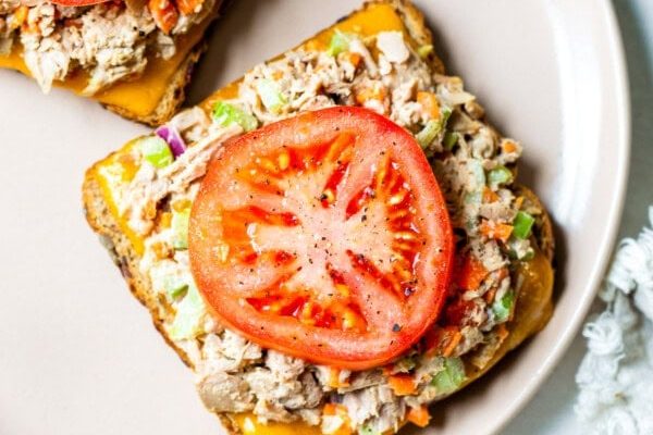 GVL Two open-faced tuna melts on a beige plate, each topped with a slice of tomato, tuna salad, and melted cheese on toasted bread. A white textured cloth is partially visible beside the plate.