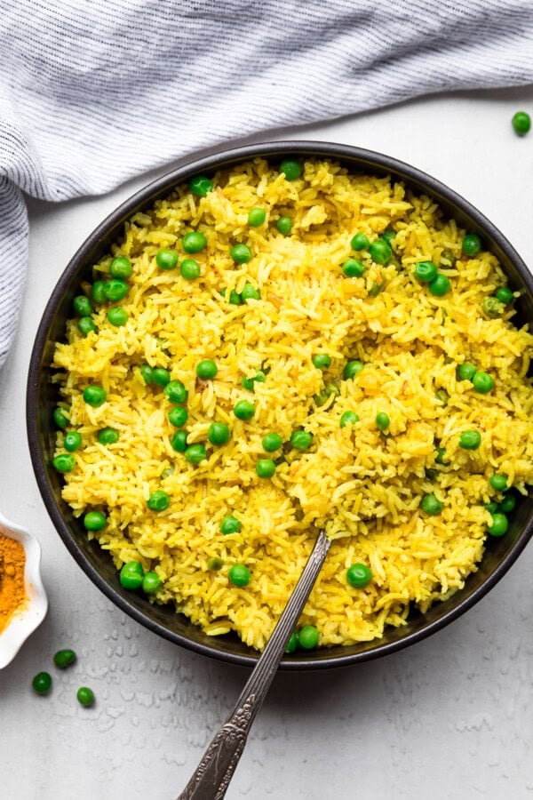 GVL A black bowl filled with vibrant Turmeric Basmati Rice and green peas, with a silver spoon resting in the rice. A gray and white striped cloth and a few scattered peas are visible nearby on the white surface.