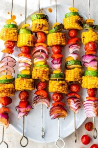 GVL Colorful vegetable skewers on metal sticks arranged on a white plate, featuring grilled corn, zucchini, cherry tomatoes, and red onion pieces—perfect for summer side dishes—with some loose tomatoes around the plate.
