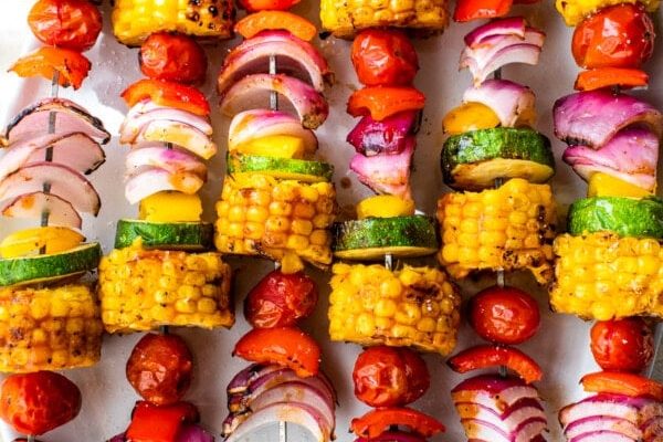 GVL Colorful vegetable skewers on metal sticks arranged on a white plate, featuring grilled corn, zucchini, cherry tomatoes, and red onion pieces—perfect for summer side dishes—with some loose tomatoes around the plate.