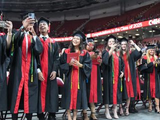 GVL A group of Wade Hampton graduates in black caps and gowns with red stoles stand smiling and celebrating, some taking selfies, at a graduation ceremony inside an arena. A banner behind them reads, Congratulations to the Class of 2025.