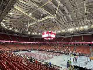 GVL Large indoor arena with red seats and a high ceiling, partially filled with people. A stage and rows of chairs are set up on the floor for Greenville County Schools high school graduations, with live bright, even lighting and a central scoreboard.