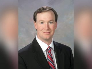 GVL Walter Wilkins, dressed in a black suit, white shirt, and red striped tie, poses for a formal headshot against a neutral grayish background, marking his retirement as 13th Circuit Court solicitor.
