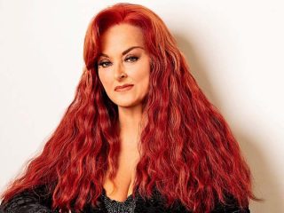 GVL A woman with long, wavy, bright red hair poses confidently against a plain light background, reminiscent of Wynonna Judd on her greatest hits tour, wearing a black outfit and subtle makeup with a slight smile.