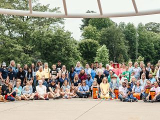 GVL A large group of people, diverse in age and appearance, pose together outdoors on a sunny day in Greenville for a Cancer Survivors Day celebration. Some are sitting, some standing, and many are smiling at the camera with trees and greenery in the background.