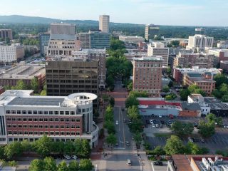 GVL Aerial view of Greenville’s city center with mid-rise office buildings, streets lined with trees, and parked cars; the downtown conference center stands out as green hills rise in the distance under a clear sky.