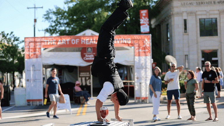 GVL A street performer does a one-handed handstand on a table with a glass of wine, while people watch and take photos at an outdoor event. A banner reading The Art of Health Living is visible, highlighting the buzz around upcoming Artisphere events.