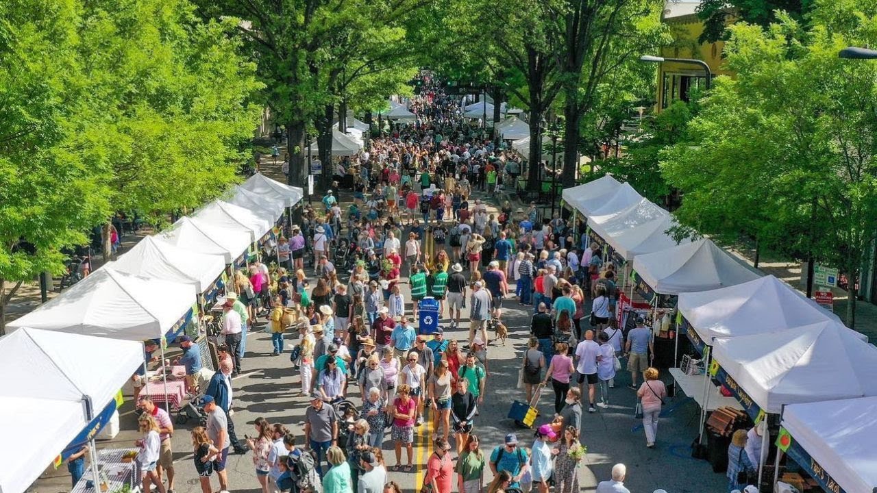 GVL A bustling outdoor street market with white vendor tents on both sides, crowded with people browsing stalls under leafy green trees on a sunny day.