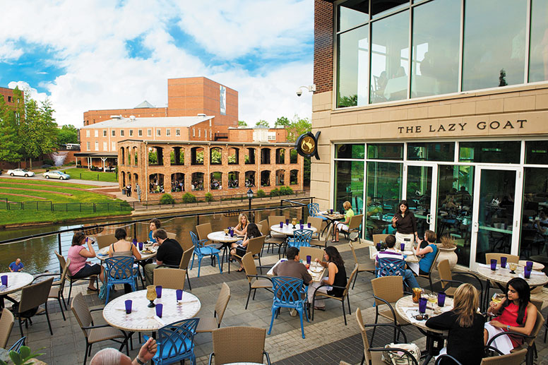 GVL People dine at outdoor tables on a patio beside a river, outside a restaurant called The Lazy Goat. The scene is bright and lively, with a historic brick building and green trees in the background.