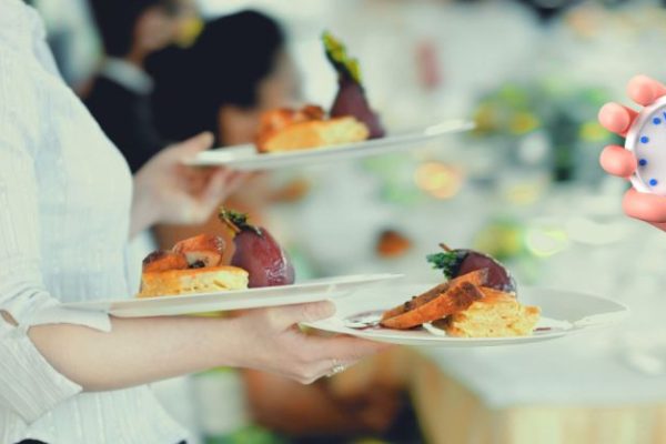 GVL A server carries plates of food at a restaurant, while a cartoon hand holding a stopwatch appears in the foreground, suggesting a focus on timing or speed of service.