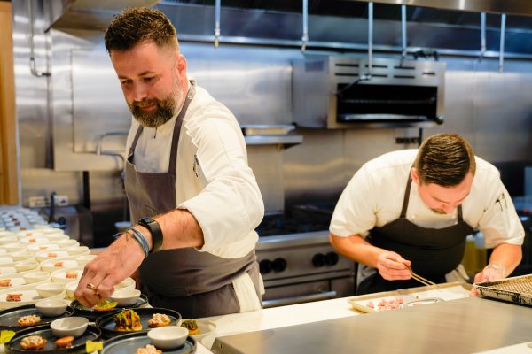 GVL Two chefs in white uniforms and aprons are carefully plating food in a professional kitchen, focusing intently on their work with various small dishes and ingredients arranged on the counter.