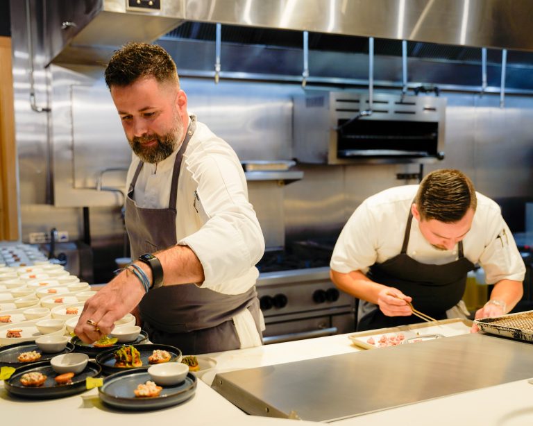 GVL Two chefs in white uniforms and aprons are carefully plating food in a professional kitchen, focusing intently on their work with various small dishes and ingredients arranged on the counter.