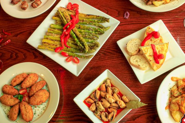 GVL A variety of tapas dishes on a wooden table, including asparagus with red peppers, bread with tortilla, mussels, croquettes, potato dishes, and a cocktail with lime and berries.