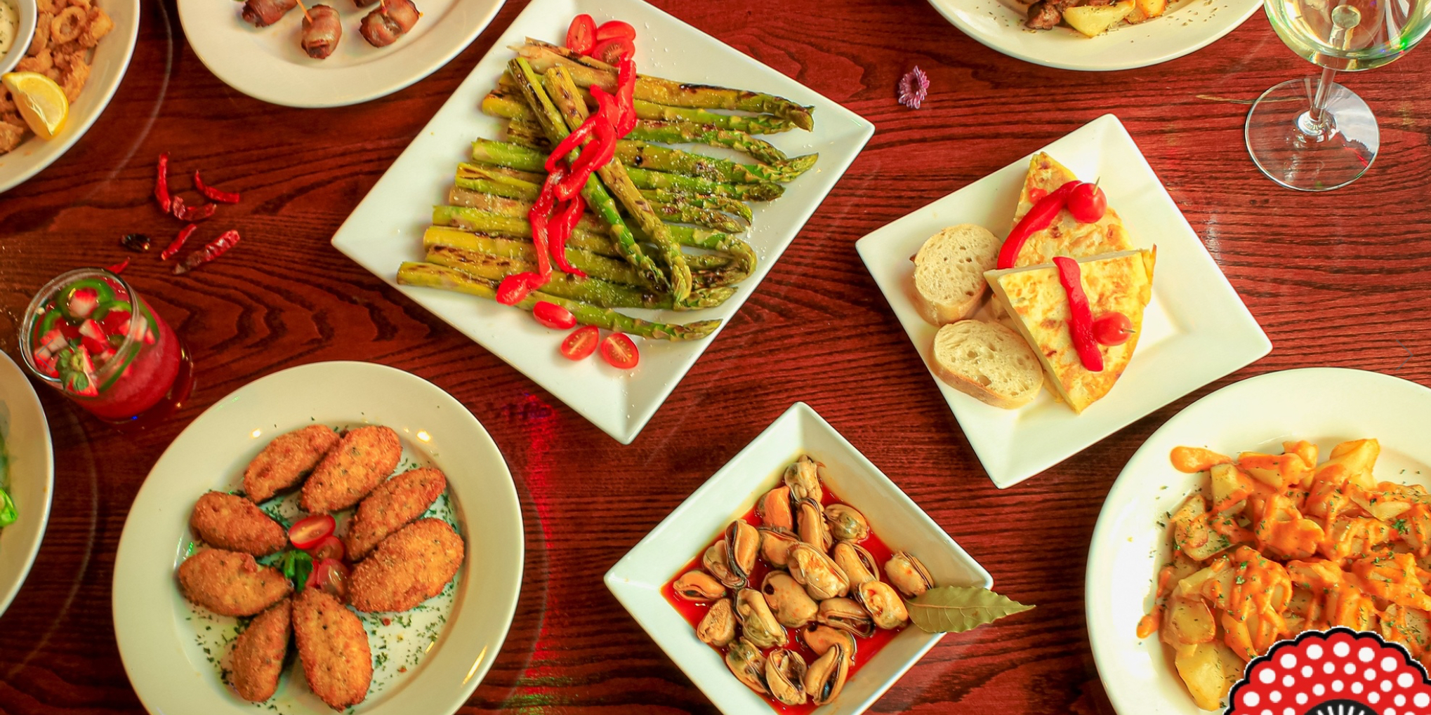 GVL A variety of tapas dishes on a wooden table, including asparagus with red peppers, bread with tortilla, mussels, croquettes, potato dishes, and a cocktail with lime and berries.
