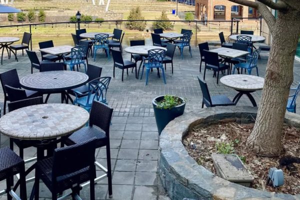 GVL Outdoor patio with empty tables and chairs on a stone-paved surface. There is a tree planted in a circular stone planter, and buildings with windows and a grassy area are visible in the background.