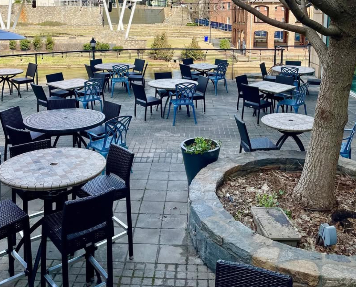 GVL Outdoor patio with empty tables and chairs on a stone-paved surface. There is a tree planted in a circular stone planter, and buildings with windows and a grassy area are visible in the background.