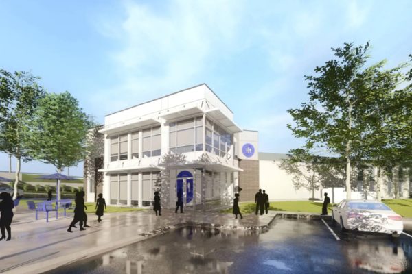 GVL A modern two-story building with large glass windows, people walking nearby, trees, parked car, and outdoor tables, all under a blue sky with clouds reflected in a puddle on the pavement.