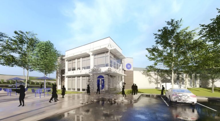 GVL A modern two-story building with large glass windows, people walking nearby, trees, parked car, and outdoor tables, all under a blue sky with clouds reflected in a puddle on the pavement.