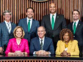 GVL Seven people pose for a formal group photo; four men stand in back wearing suits and ties, while two women and one man, possibly City Council members discussing the $291M Greenville budget, sit in front. The background features vertical wooden panels.