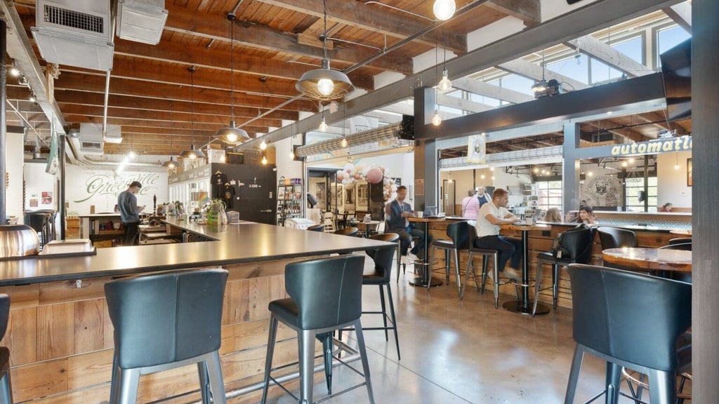GVL Open, modern cafe with wooden ceilings, exposed beams, and large windows. People sit at high tables and bar seating, some working on laptops and others talking. Warm lighting and a relaxed atmosphere.