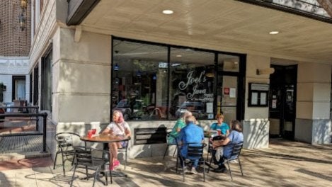 GVL Four people sit at two outdoor tables in front of a café called Joel’s Java on a sunny day. The café has large windows, and people can be seen inside. The sidewalk and tables are shaded by the building.