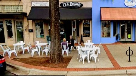 GVL Outdoor seating area in front of Old Europe Coffee & Pastries café, with several white tables and chairs on the sidewalk. Two people are sitting at one table under a tree, talking.
