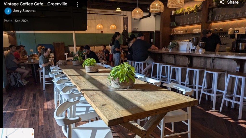 GVL A cozy coffee shop with wooden tables, white chairs, hanging wicker lights, and potted plants. People sit at the bar and tables, enjoying drinks. Natural light streams in through the windows, creating a warm atmosphere.