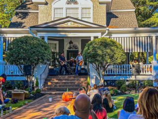 GVL A small band performs on the porch of a large, classic house during Hampton-Pinckney Porchfest, while people sit on the lawn enjoying the outdoor concert—a true showcase of heritage tourism on a sunny day surrounded by trees, shrubs, and pumpkins.