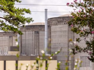 GVL Large concrete cylindrical structures of a Duke Energy nuclear power plant are partially obscured by trees and foliage, with a cloudy sky in the background. Thin vertical pipes extend above the buildings, hinting at increased power usage during a heat wave.