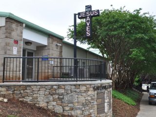 GVL A building with a stone wall and metal railing features a large black cross sign reading JESUS SAVES in white letters. Trees and greenery thrive nearby, with Miracle Hill known for providing severe weather shelters to those in need.