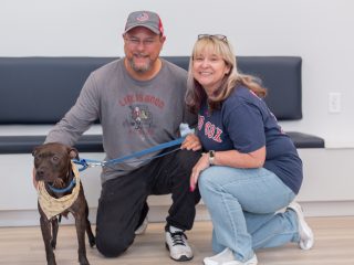 GVL A smiling man and woman kneel next to a brown dog wearing a bandana and blue leash, celebrating their recent dog adoption. They are indoors, in front of a bench and white wall at the Greenville County animal hospital.