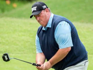 GVL A man wearing a black cap, blue shirt, and navy vest is playing golf on a grassy course during the BMW Charity Pro-Am, concentrating as he swings a golf club.