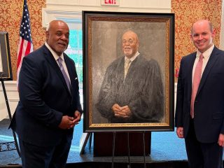 GVL Two men in suits stand on either side of a framed portrait of a Greenville judge in robes, posing in a formal room with patterned wallpaper and an American flag in the background, celebrating the Order of the Palmetto.