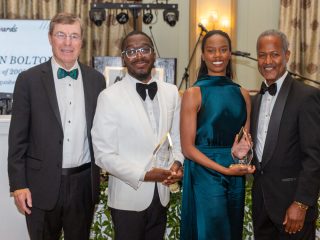 GVL Four people dressed formally stand together at the SC Governor’s School 25th anniversary gala. Two individuals in the center hold clear glass awards, with a sign and musical equipment visible in the background of this fundraising event.