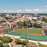 GVL Aerial view of Greenville’s baseball stadium surrounded by trees and buildings, with a city skyline visible in the background under a partly cloudy sky, as seen on CBS Evening News.