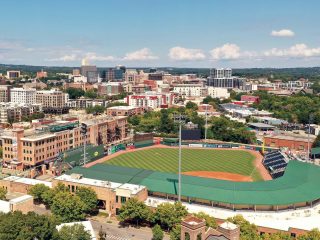 GVL Aerial view of Greenville’s baseball stadium surrounded by trees and buildings, with a city skyline visible in the background under a partly cloudy sky, as seen on CBS Evening News.