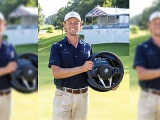 GVL A smiling golfer, one of the winners at the BMW Charity Pro-Am, stands on a golf course holding a BMW steering wheel as a trophy, with a clubhouse and trees in the background.