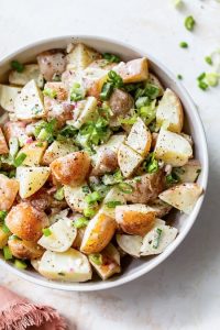 GVL A bowl of creamy Red Potato Salad with chopped green onions, red onions, and black pepper on top, served in a white bowl on a light surface.