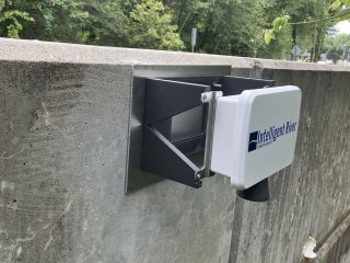 GVL A small electronic device labeled North Virginia Water, part of a water-sensor system, is mounted on a concrete wall outdoors, with trees and greenery visible in the background.