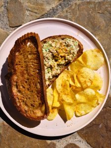 GVL A plate with a toasted Chickpea Salad Sandwich filled with an egg salad mixture, served alongside a pile of potato chips. The plate is set on a stone surface.