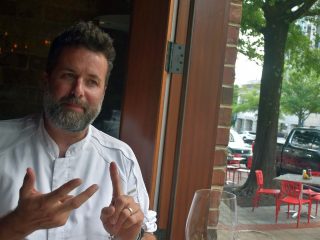 GVL A man with a beard and chef’s jacket sits by a window in a restaurant, gesturing with his hands. Outside, red chairs and tables are visible on the sidewalk near City Juice and parked cars.