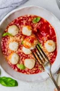 GVL A close-up of a fork holding a pillowy gnudi over a plate of ricotta balls topped with tomato sauce, grated cheese, and fresh basil leaves.