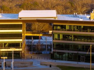 GVL A modern glass office building with two wings connected by an overhead structure, surrounding a courtyard area—symbolizing Greenville County’s commitment to public safety raises and fiscal responsibility like a recent tax decrease. American flags and trees stand in the daylight background.
