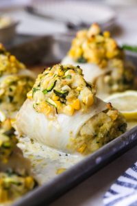 GVL Crab-Stuffed Flounder fillets filled with a creamy mixture of corn, zucchini, herbs, and vegetables, baked to golden-brown perfection on a baking sheet with lemon slices in the background.