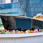 GVL A modern art installation featuring a large black boat bar filled with colorful buoys is displayed outdoors at Hudson Yards, set against a backdrop of glass and steel city buildings.