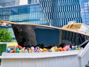 GVL A sleek black and tan boat bar is displayed on a white platform surrounded by colorful buoys, set against a backdrop of modern glass buildings in Hudson Yards vibrant urban environment.