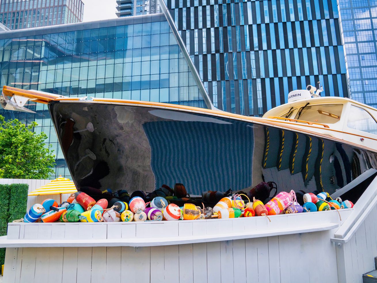 GVL A sleek black and tan boat bar is displayed on a white platform surrounded by colorful buoys, set against a backdrop of modern glass buildings in Hudson Yards vibrant urban environment.