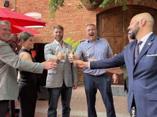 GVL Five people stand outdoors in a semicircle at Soby’s, smiling and raising glasses in a toast. Dressed in business or smart casual attire, they enjoy the ambiance of one of the top SC restaurants, with red umbrellas and a brick building behind them.