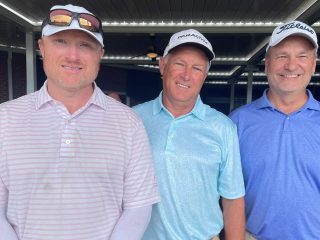 GVL Three men in golf attire stand side by side, smiling at the camera during a Laurens Electric golf tournament. They are wearing collared shirts and caps, outdoors under a covered area with ceiling lights. The event raises $21K.