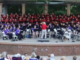 GVL A large choir and orchestra perform on an outdoor stage at Furman. The conductor faces musicians in purple shirts, with singers in red and black filling the tiered rows behind them. Audience members seated in front enjoy this patriotic concert.